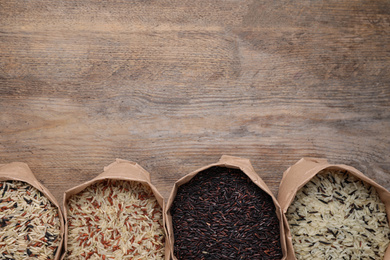 Photo of Different types of brown and polished rice in paper bags on wooden table, flat lay. Space for text