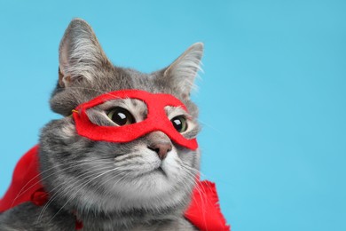 Photo of Adorable cat in red superhero cape and mask on light blue background, closeup. Space for text