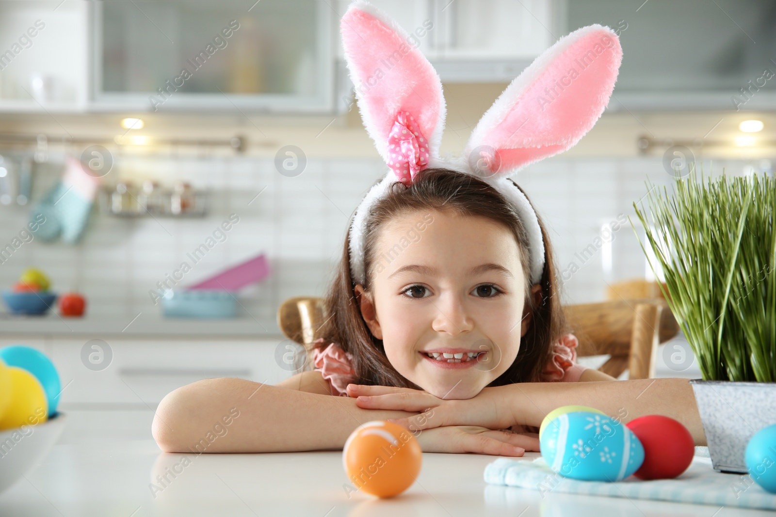 Photo of Cute little girl with bunny ears headband and painted Easter eggs sitting at table in kitchen, space for text