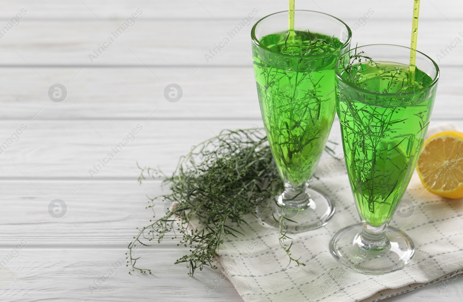 Photo of Glasses of homemade refreshing tarragon drink, sprigs and lemon on white wooden table, space for text