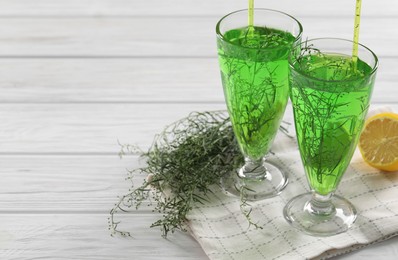 Photo of Glasses of homemade refreshing tarragon drink, sprigs and lemon on white wooden table, space for text