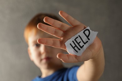 Photo of Little boy holding piecepaper with word Help against light grey background, focus on hand. Domestic violence concept