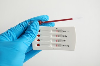 Photo of Doctor holding disposable express test for hepatitis and pipette with blood on white background, closeup