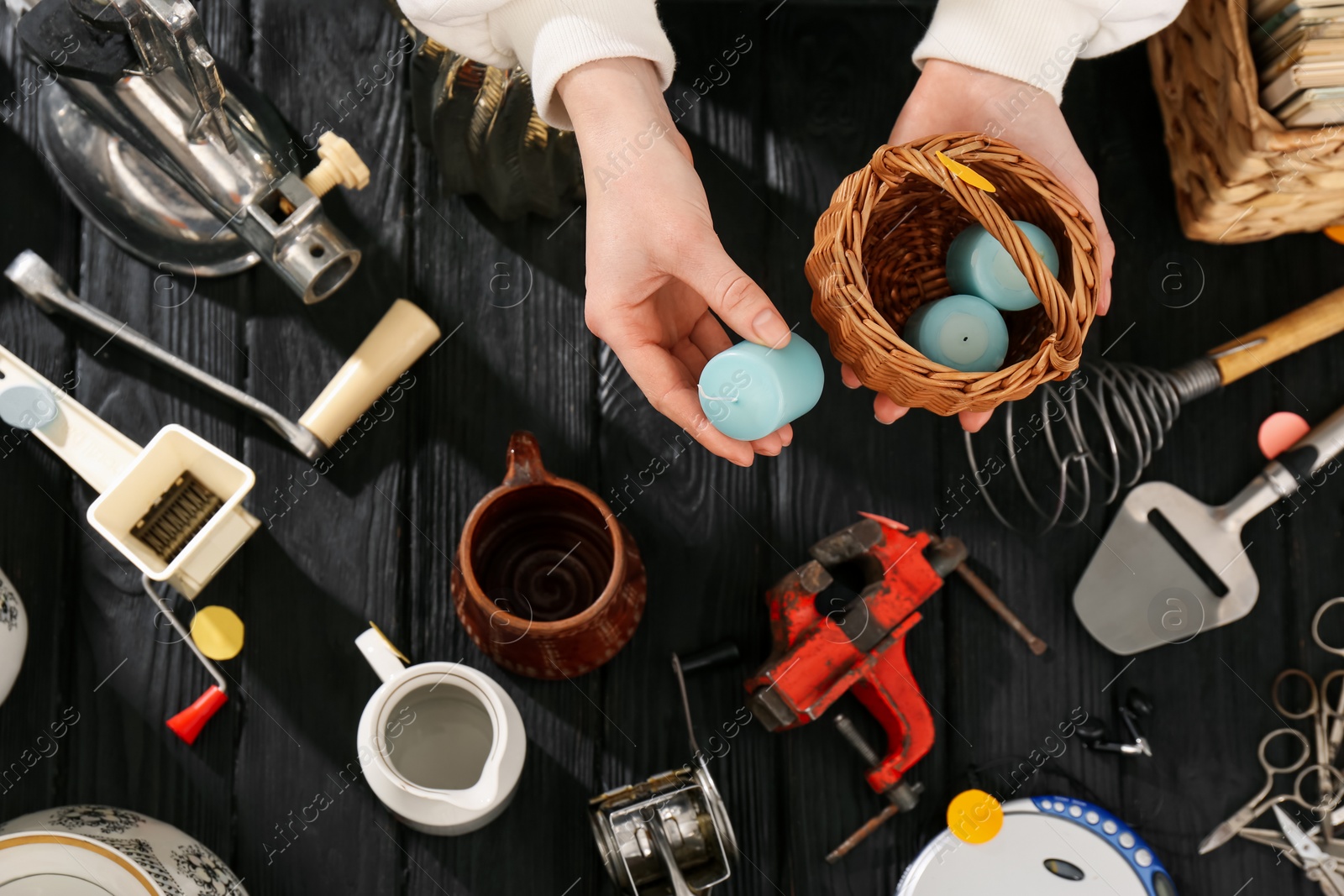 Photo of Woman putting candle into wicker basket above black wooden table with different stuff, top view. Garage sale