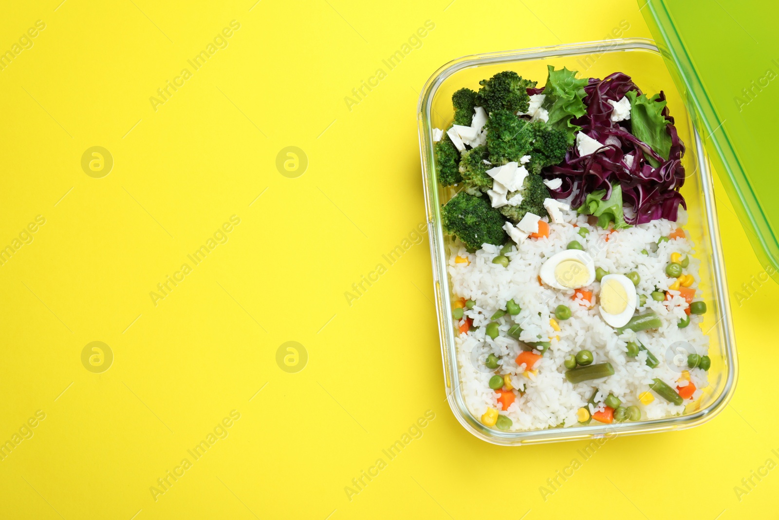 Photo of Tasty rice with vegetables and salad in glass container on yellow background, top view. Space for text