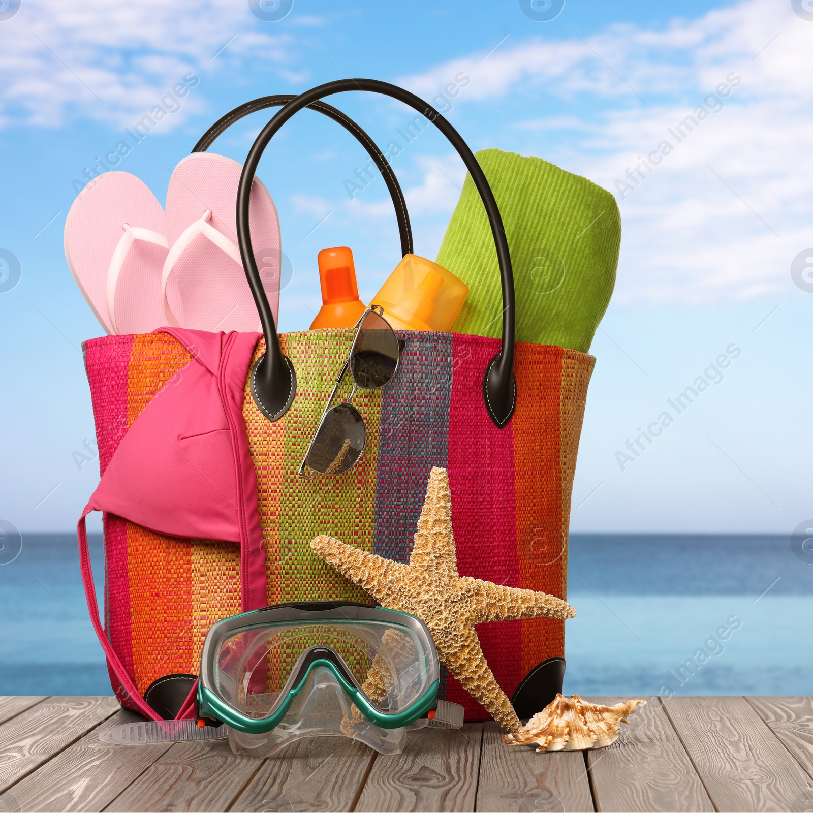 Image of Stylish bag with beach accessories on wooden surface near seashore 