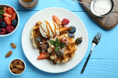 Photo of Delicious Belgian waffles with ice cream, berries and caramel sauce served on light blue wooden table, flat lay