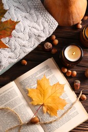 Book with autumn leaf as bookmark, acorns, scented candles and warm sweaters on wooden table, flat lay