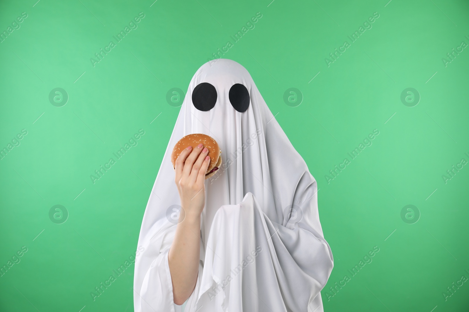 Photo of Creepy ghost. Person in white sheet with tasty hamburger on green background