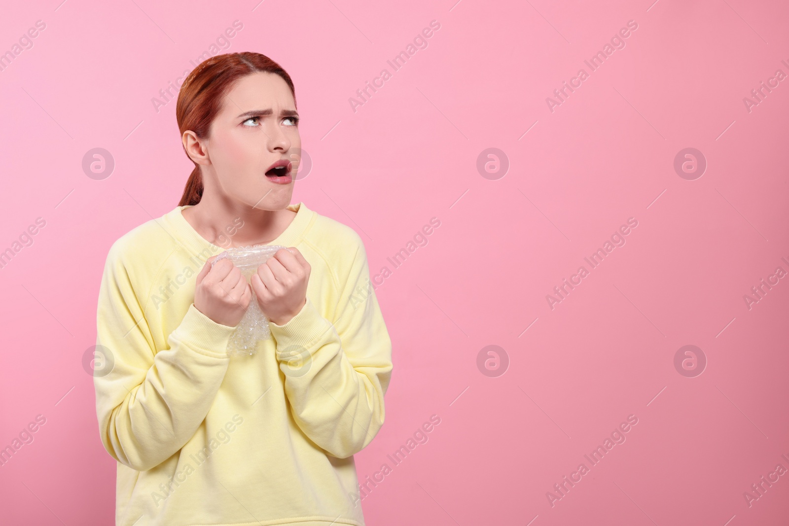 Photo of Angry woman popping bubble wrap on pink background, space for text. Stress relief