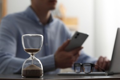 Photo of Hourglass with flowing sand on desk. Man using smartphone while working on laptop indoors, selective focus