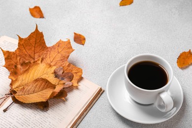 Cup of hot drink, book and autumn leaves on light grey textured table