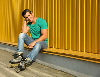 Photo of Handsome young man with inline roller skates sitting near yellow building, space for text