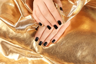 Photo of Woman showing black manicure on golden fabric, closeup. Nail polish trends