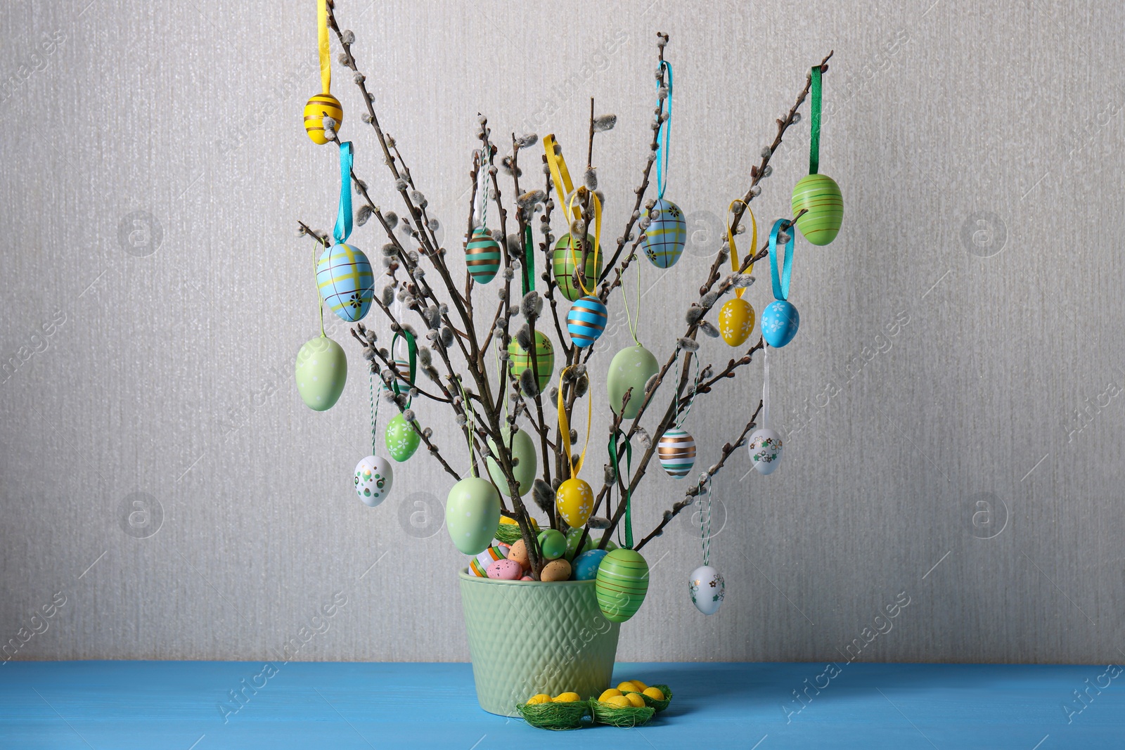 Photo of Beautiful willow branches with painted eggs in pot on light blue table. Easter decor