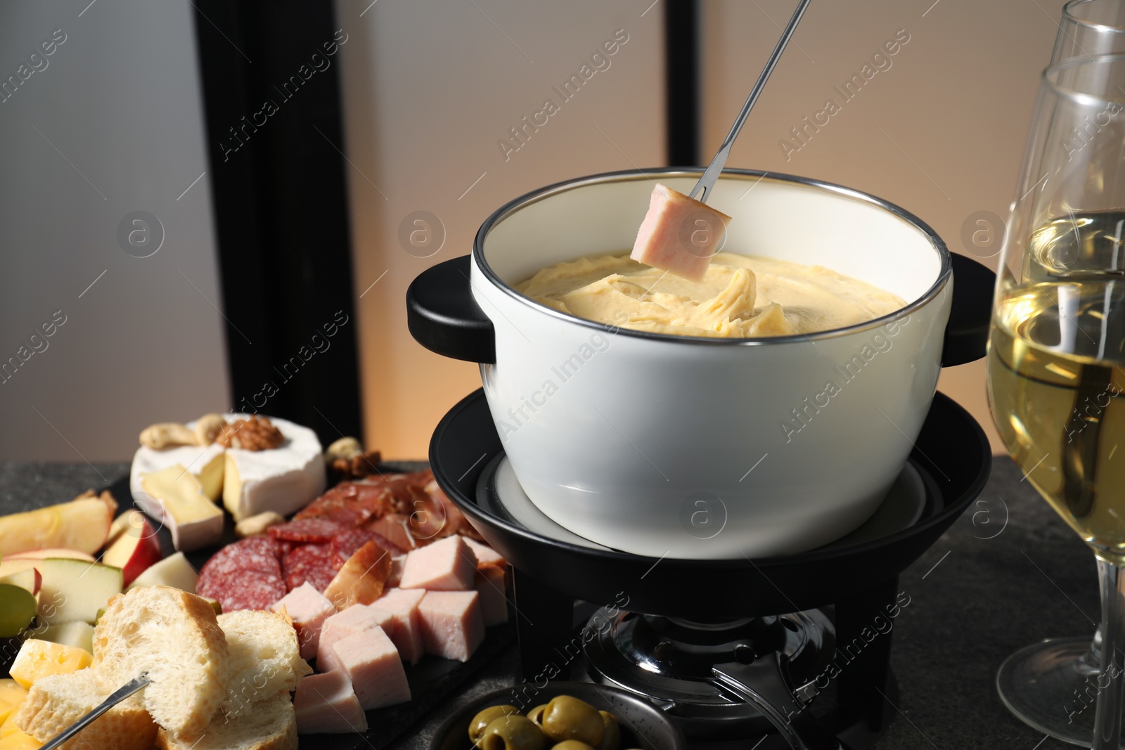 Photo of Dipping piece of ham into fondue pot with melted cheese at grey table with wine and snacks, closeup