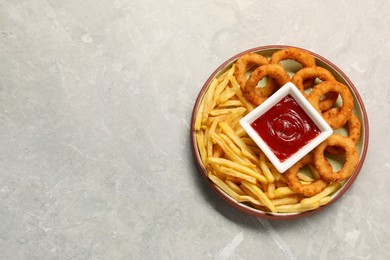 Plate with tasty ketchup, fries and onion rings on light grey table, top view. Space for text