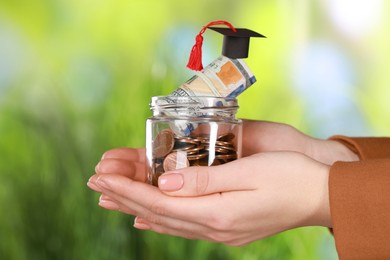 Photo of Woman holding glass jar with coins, dollar banknotes and graduation cap against blurred background, closeup. Scholarship concept