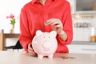 Photo of Woman putting coin into piggy bank at table, closeup. Saving money