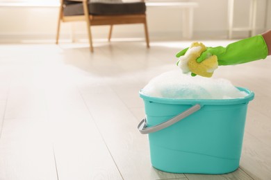 Photo of Woman holding sponge with foam over bucket indoors, closeup. Cleaning supplies