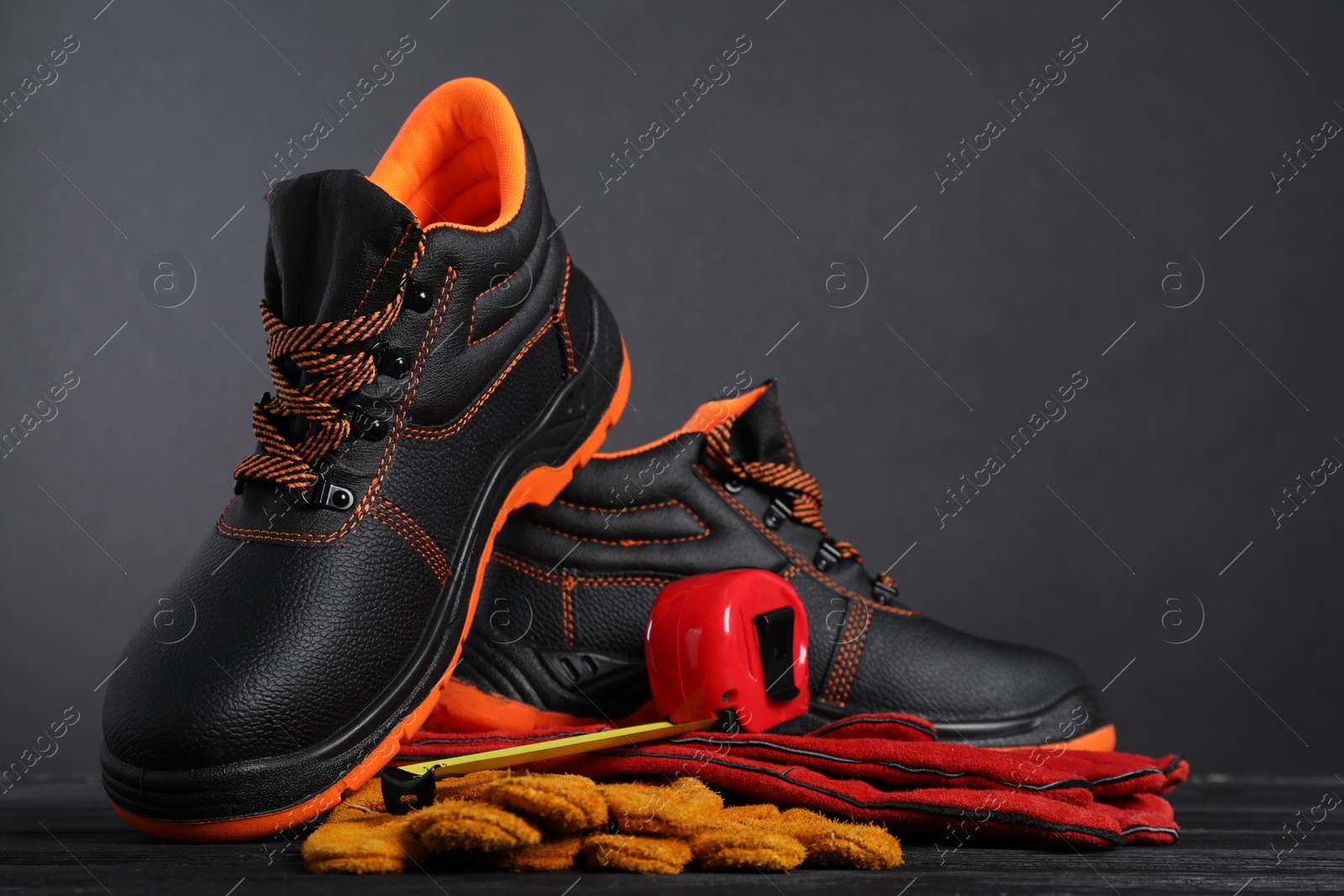 Photo of Pair of working boots, protective gloves and tape measure on black wooden surface against gray background, space for text