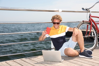 Attractive man with laptop and bike near sea on sunny day, space for text