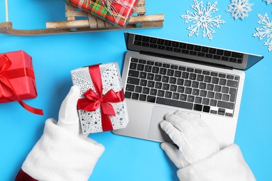Photo of Santa Claus using laptop, closeup. Gift boxes and Christmas decor on light blue background, top view