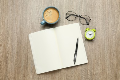 Open notebook, glasses, alarm clock, pen and cup of coffee on wooden table, flat lay