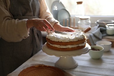 Photo of Woman smearing sponge cake with cream at white wooden table in kitchen, closeup