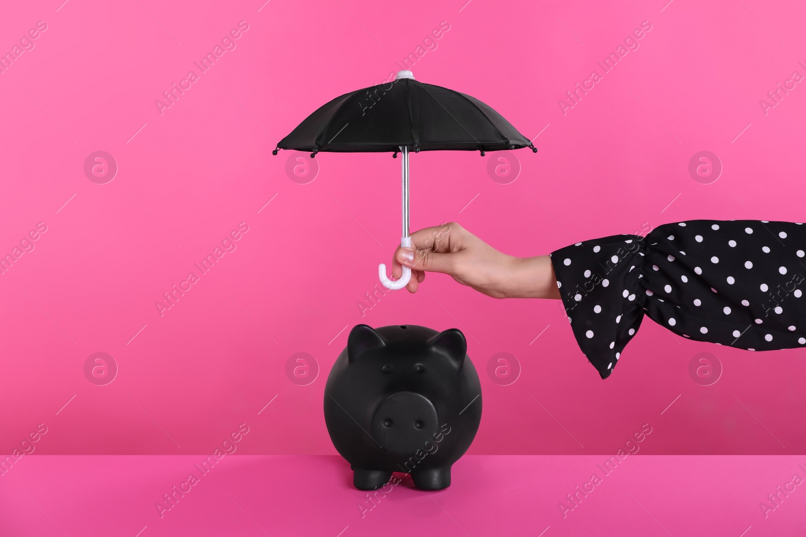 Photo of Woman holding small umbrella over piggy bank on pink background, closeup