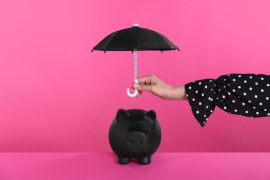 Photo of Woman holding small umbrella over piggy bank on pink background, closeup