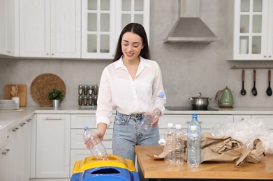 Photo of Garbage sorting. Smiling woman throwing plastic bottle into trash bin in kitchen