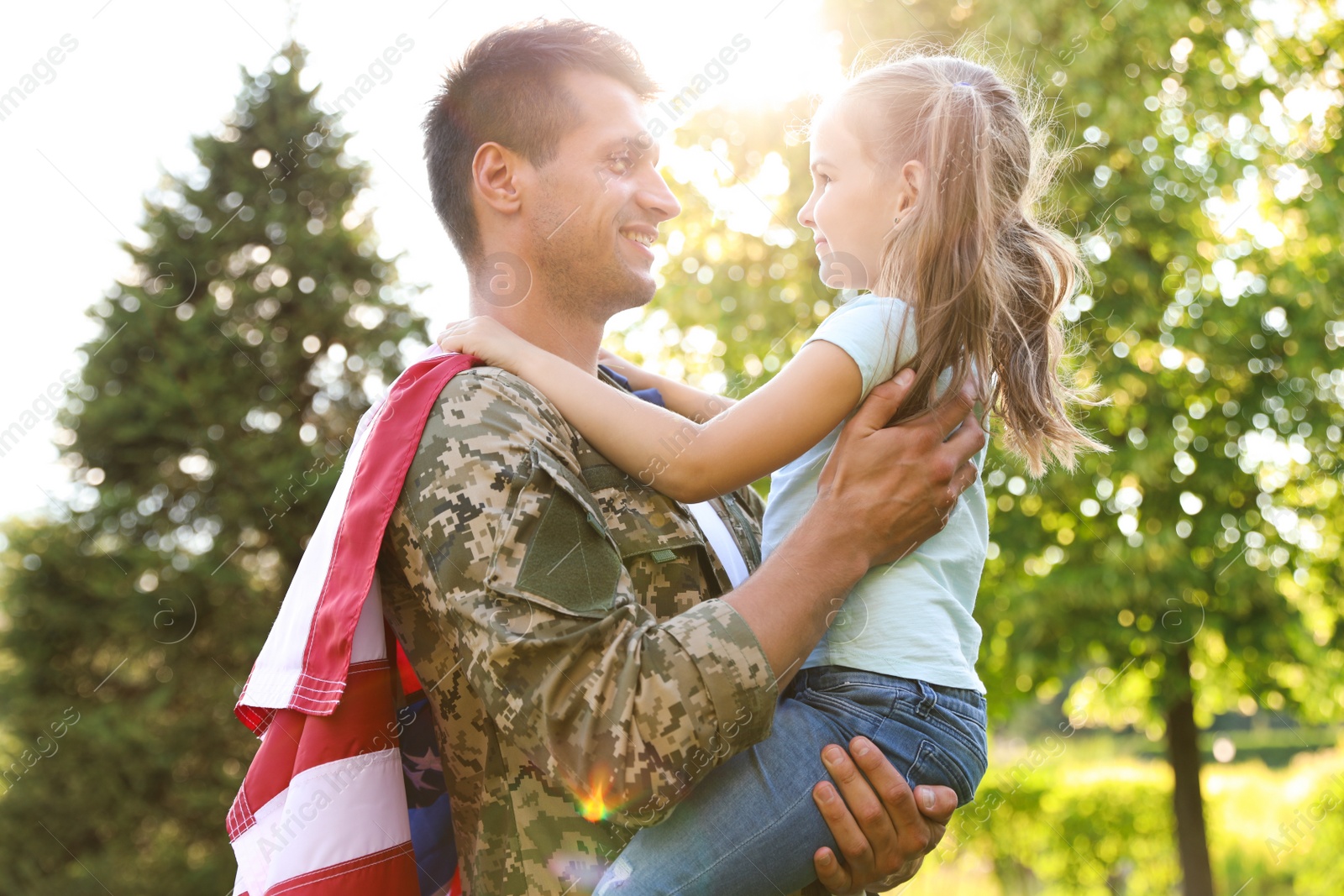 Photo of Father in military uniform with American flag holding his little daughter at green park