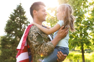 Photo of Father in military uniform with American flag holding his little daughter at green park