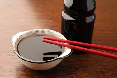 Photo of Bottle, bowl with soy sauce and chopsticks on wooden table, closeup