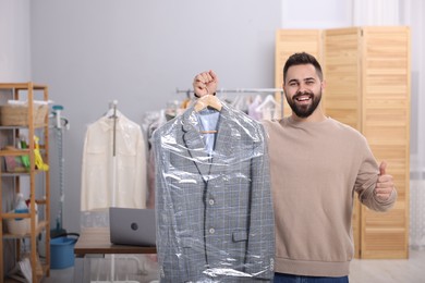Photo of Dry-cleaning service. Happy man holding hanger with jacket in plastic bag and showing thumb up indoors, space for text