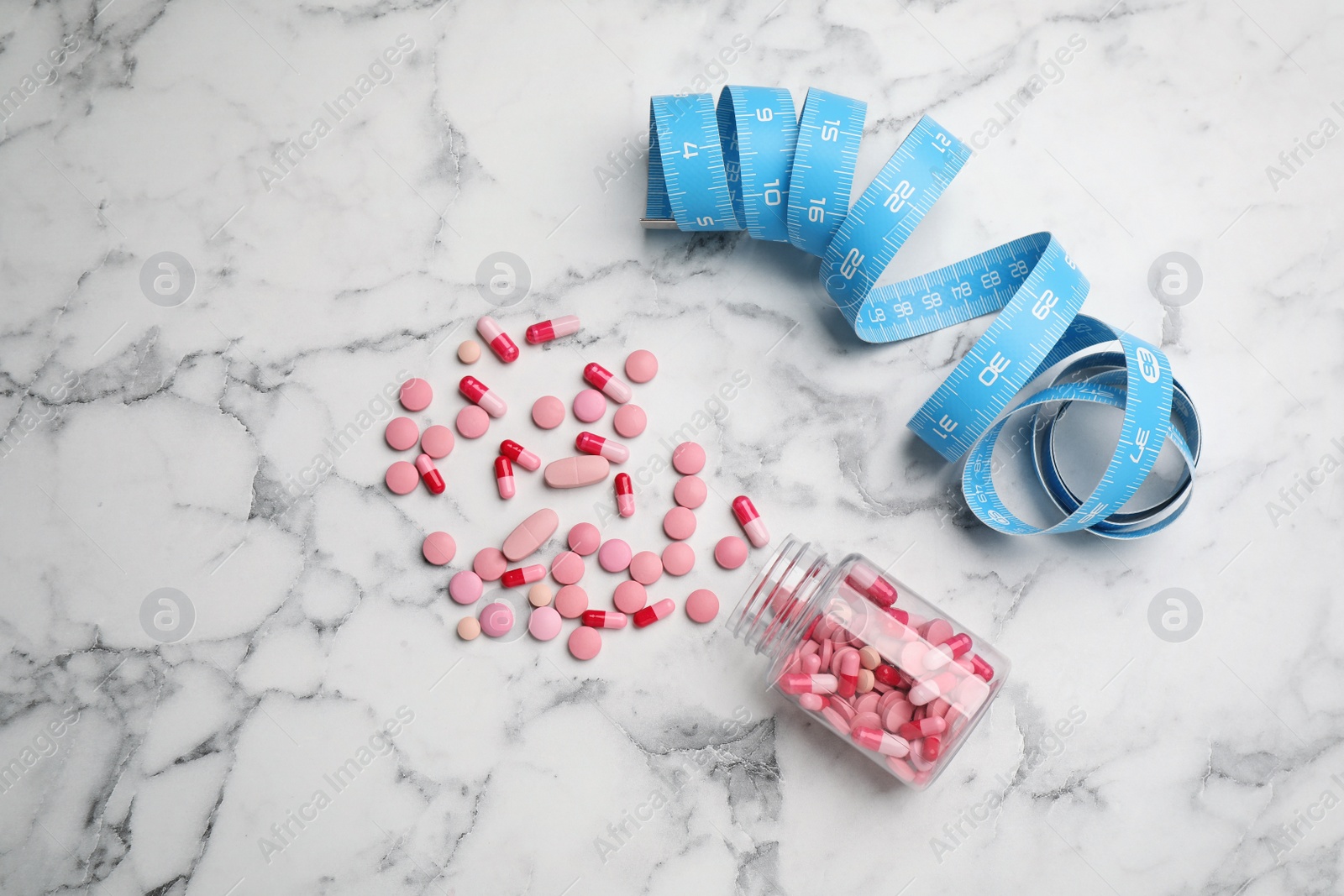 Photo of Jar of weight loss pills and measuring tape on white marble table, flat lay