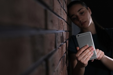 Sad woman with mobile phone near brick wall, space for text. Loneliness concept
