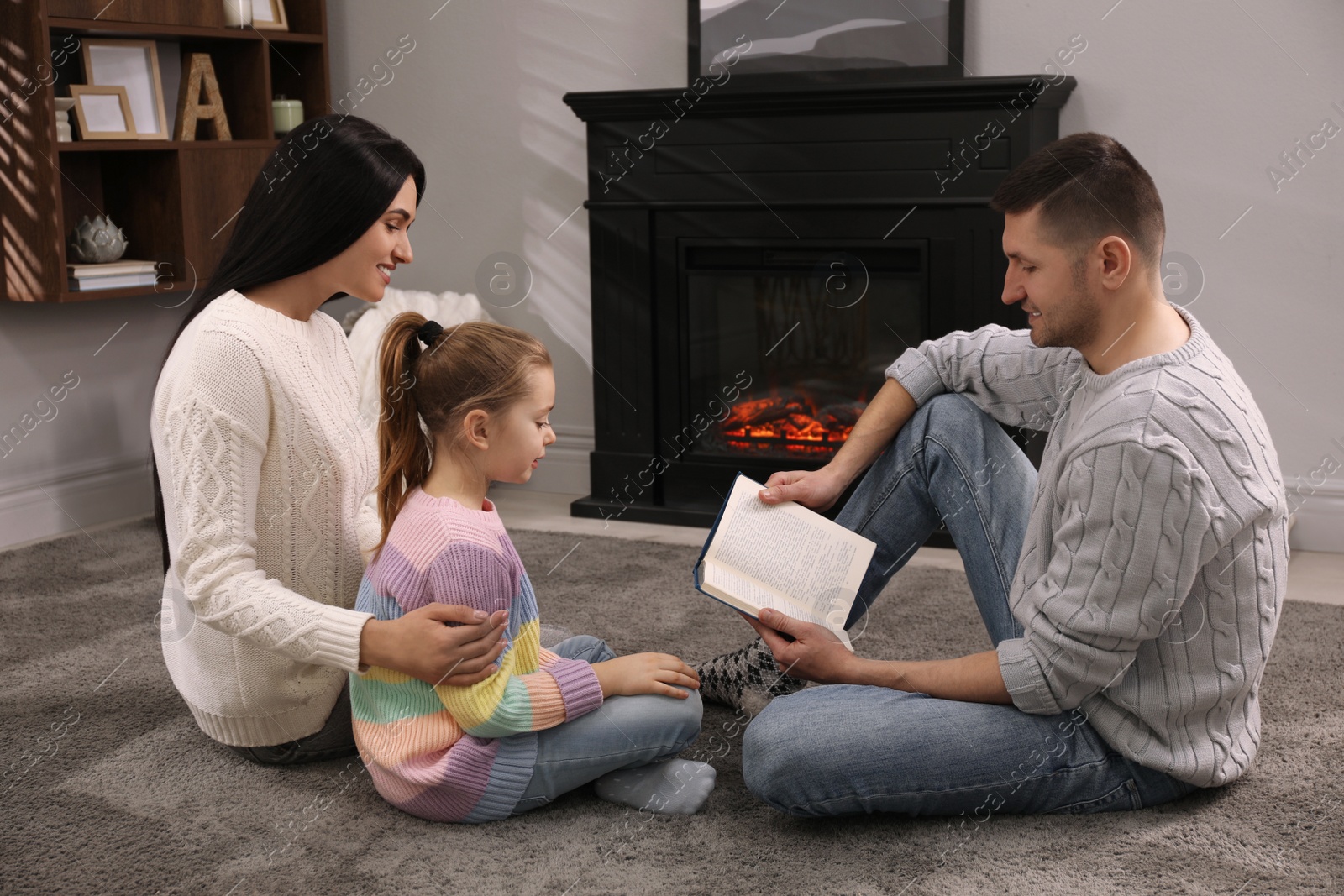 Photo of Happy family reading book together on floor near fireplace at home