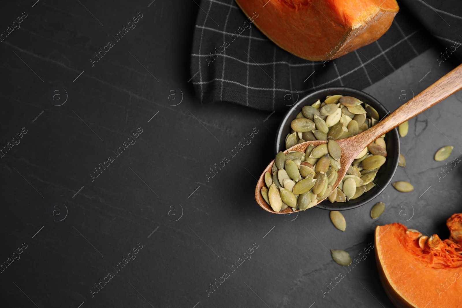Photo of Bowl with seeds, wooden spoon and cut pumpkin on black table, flat lay. Space for text