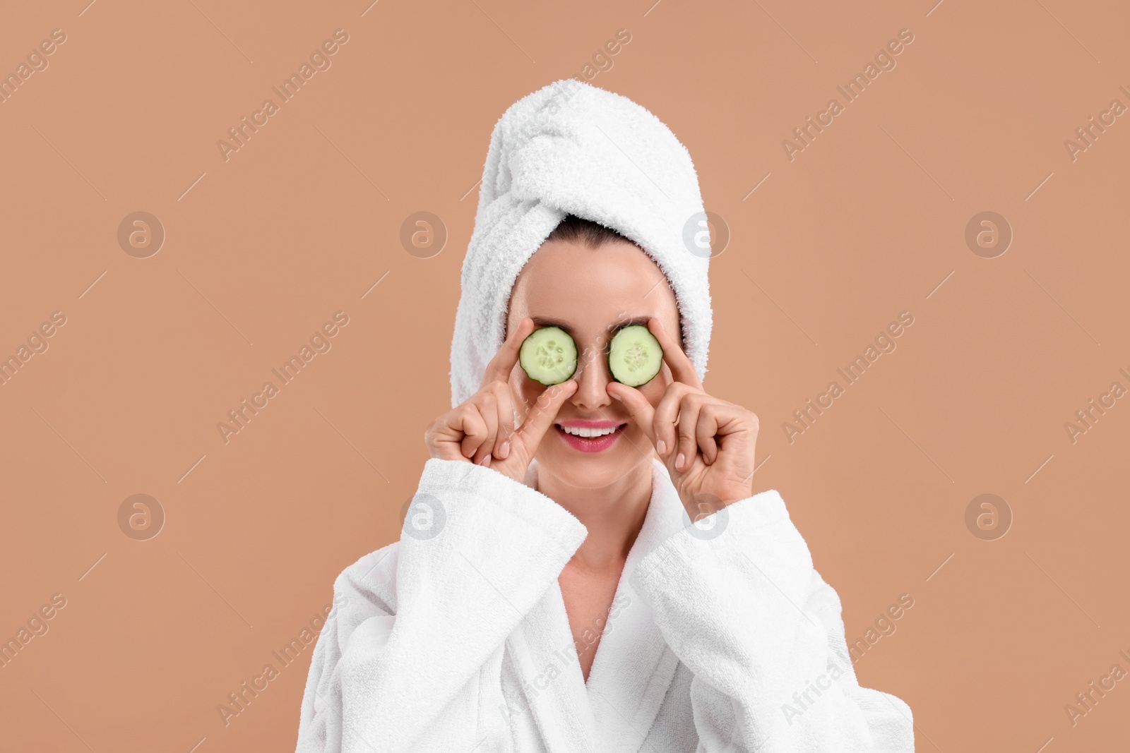 Photo of Beautiful woman in bathrobe covering eyes with pieces of cucumber on beige background