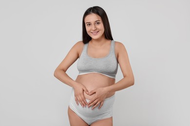 Photo of Beautiful pregnant woman in comfortable maternity underwear making heart with hands on her belly against grey background
