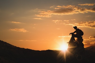 Image of Silhouette of man with backpack sitting on peak in mountains at sunset. Space for text