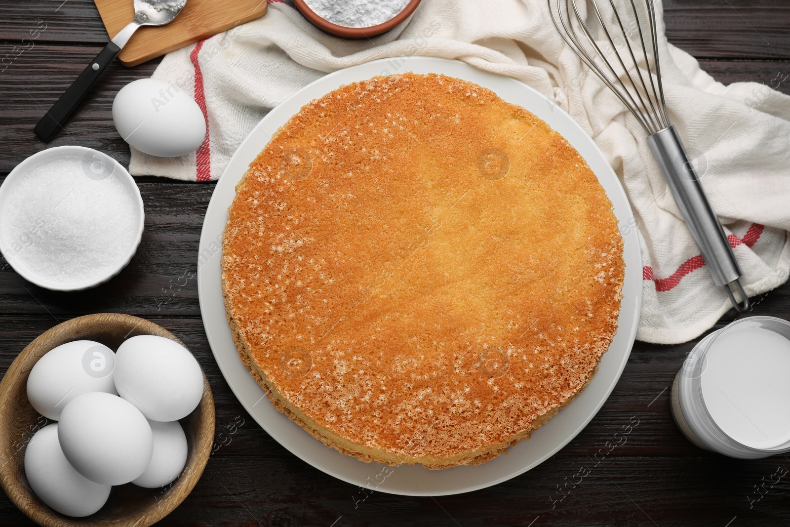 Photo of Plate with delicious sponge cake and ingredients on dark wooden table, flat lay