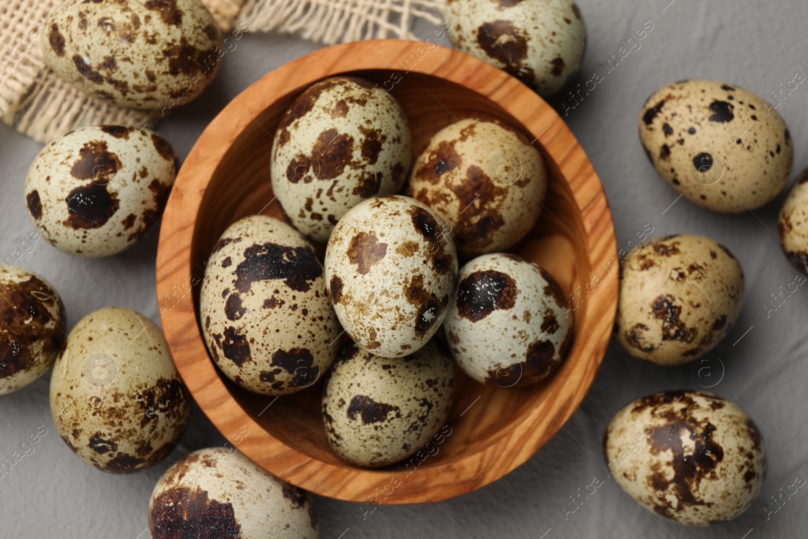 Photo of Wooden bowl and many speckled quail eggs on grey table, flat lay