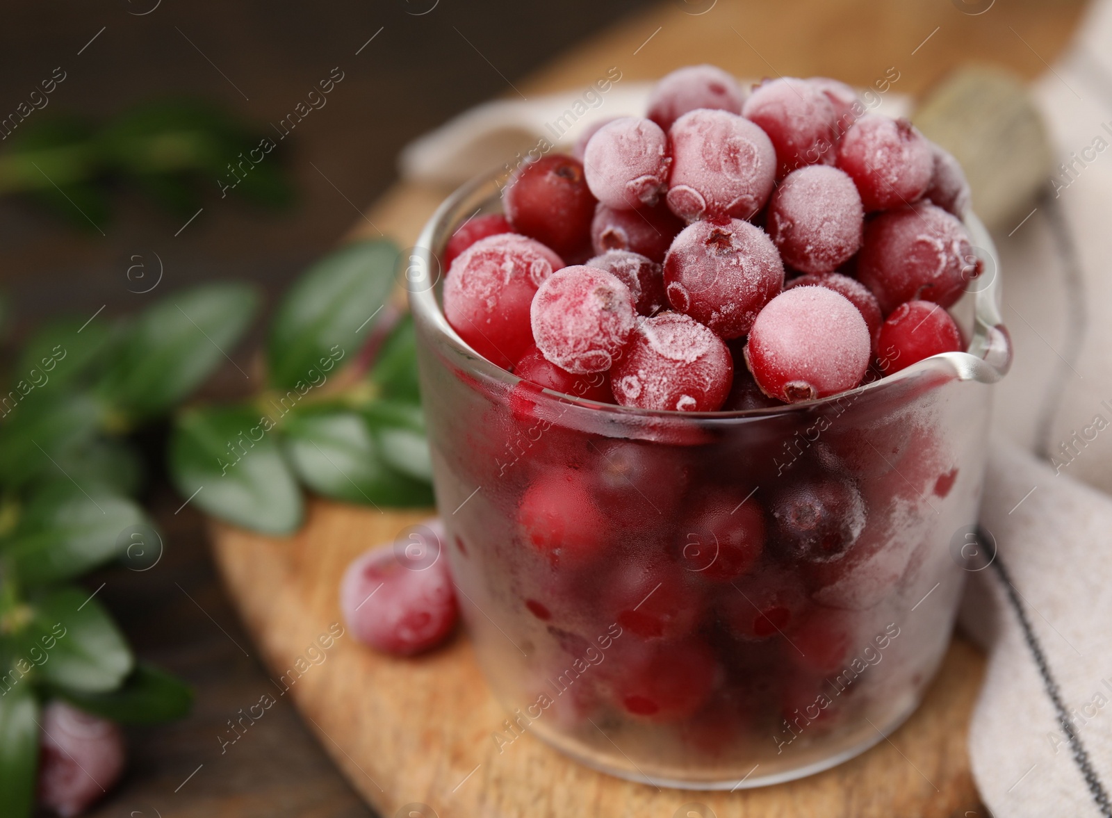 Photo of Frozen red cranberries in glass pot and green leaves on table, closeup