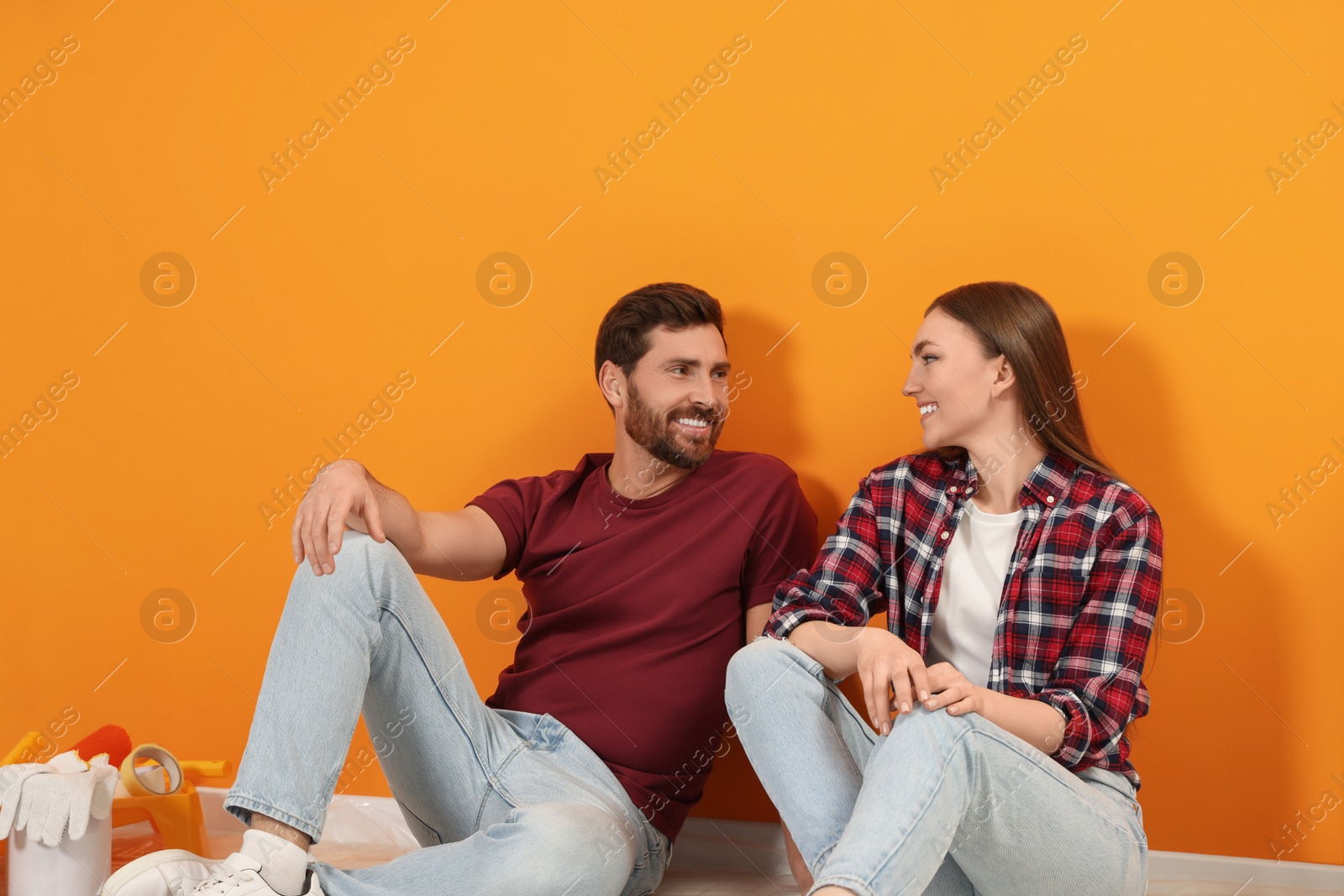Photo of Happy designers sitting on floor with painting equipment near freshly painted orange wall indoors
