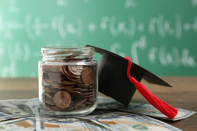 Photo of Scholarship concept. Glass jar with coins, graduation cap and dollar banknotes on wooden table