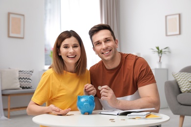 Photo of Couple putting coins into piggy bank at table in living room. Saving money