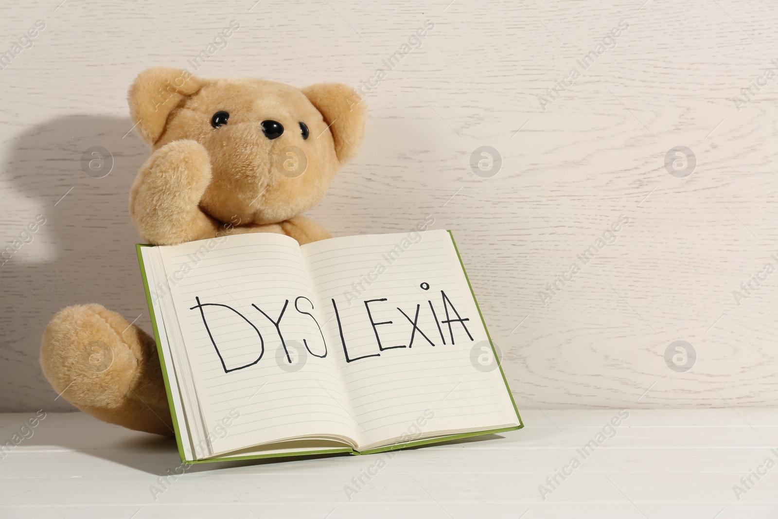 Photo of Notebook with word Dyslexia and teddy bear on white table near wooden wall, space for text